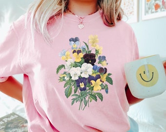May include: A pink t-shirt with a floral print of a bouquet of purple, yellow, and white pansies. The text "Blossom" is printed on the bottom right corner of the shirt.