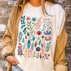 May include: A white t-shirt with a colorful floral print featuring various flowers, leaves, and stems in shades of pink, blue, green, orange, and purple.