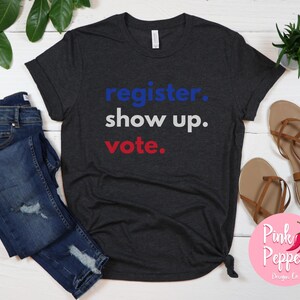 May include: A dark heather grey t-shirt with the text "register. show up. vote." in blue, white, and red lettering.