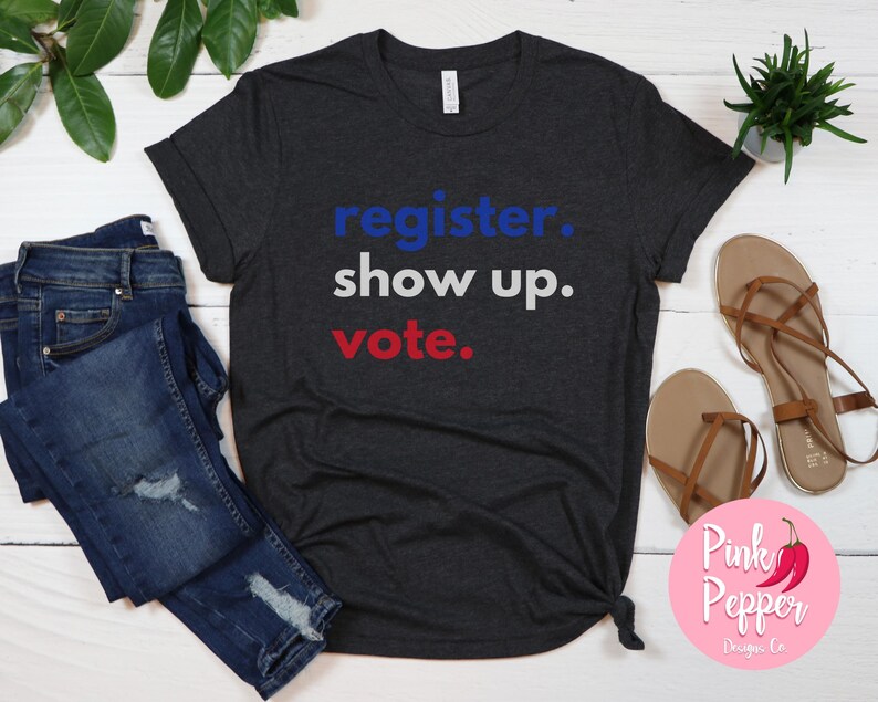 May include: A dark heather grey t-shirt with the text "register. show up. vote." in blue, white, and red lettering.
