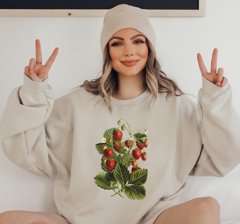 May include: A beige sweatshirt with a vintage illustration of a strawberry plant with red strawberries and green leaves.