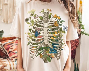 May include: Ivory-colored t-shirt featuring a detailed illustration of a human rib cage filled with colorful flowers and butterflies. The design includes various blooms and foliage, with a few butterflies. The word "ivory" is printed at the bottom.