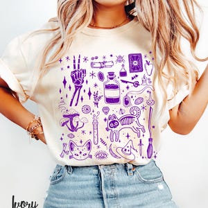 May include: A white t-shirt with a purple design of various witchy symbols, including a cat, a potion bottle, a skull, a crystal, a mushroom, a broom, and a witch's hat.