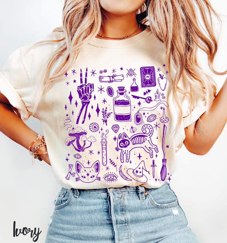 May include: A white t-shirt with a purple design of various witchy symbols, including a cat, a potion bottle, a skull, a crystal, a mushroom, a broom, and a witch's hat.