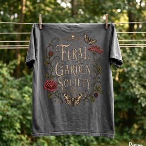 May include: Dark gray t-shirt with the words "Feral Garden Society" in a vintage font, surrounded by floral and butterfly illustrations. The shirt is hanging on a clothesline, showcasing a nature-inspired design.
