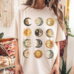 May include: A cream-colored t-shirt with a graphic print of twelve different sun and moon phases in gold, blue, and black.