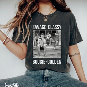 Puede incluir: Una camiseta gris oscuro con una foto en blanco y negro de cuatro personas sentadas en un porche. El texto "SAVAGE &middot; CLASSY &middot;  BOOGIE &middot; GOLDEN" est&aacute; impreso en la camiseta.