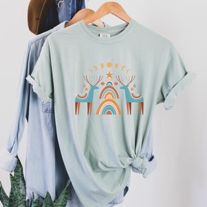 Puede incluir: Una camiseta verde claro con un dise&ntilde;o gr&aacute;fico de dos renos estilizados de pie bajo un arco&iacute;ris con estrellas y lunas. Los renos son azules y marrones, y el arco&iacute;ris es naranja, amarillo y rojo.