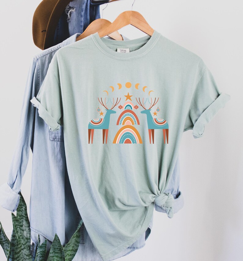 Puede incluir: Una camiseta verde claro con un dise&ntilde;o gr&aacute;fico de dos renos estilizados de pie bajo un arco&iacute;ris con estrellas y lunas. Los renos son azules y marrones, y el arco&iacute;ris es naranja, amarillo y rojo.