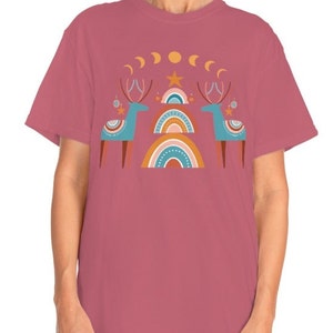 Puede incluir: Una camiseta rosa con un dise&ntilde;o gr&aacute;fico de dos renos estilizados de pie frente a un arco&iacute;ris con una estrella y lunas crecientes arriba.
