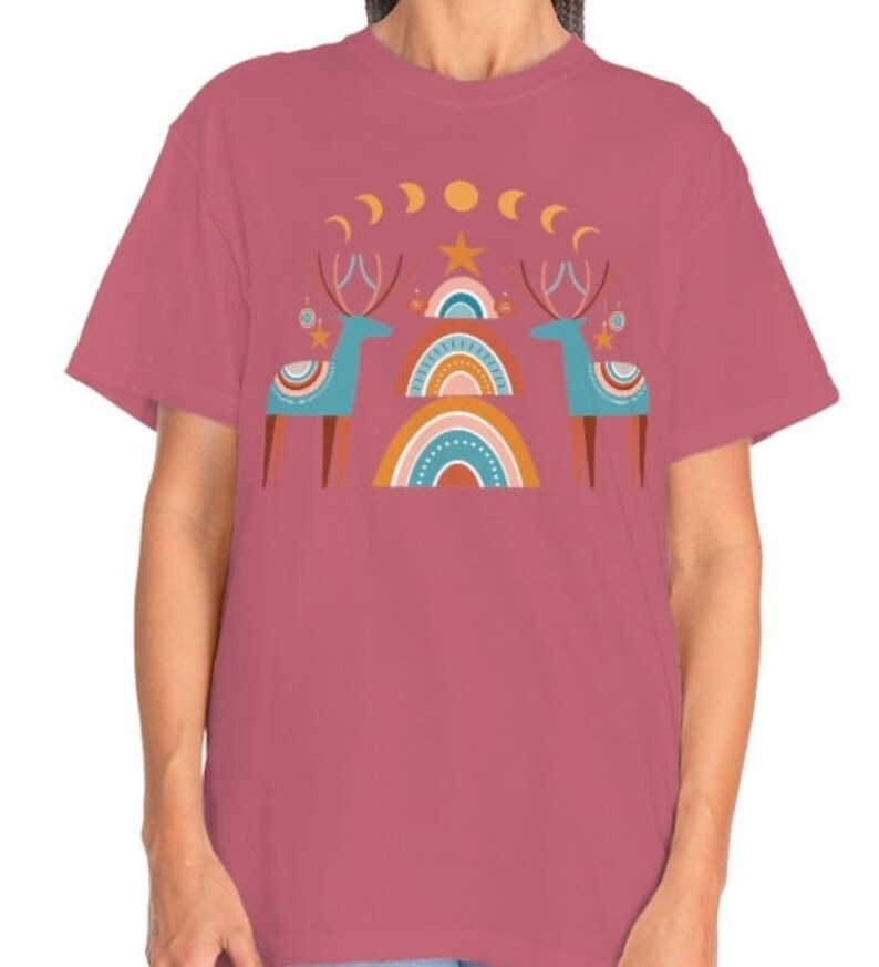 Puede incluir: Una camiseta rosa con un dise&ntilde;o gr&aacute;fico de dos renos estilizados de pie frente a un arco&iacute;ris con una estrella y lunas crecientes arriba.