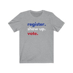 May include: A heather grey t-shirt with the text "register. show up. vote." in blue and red lettering.