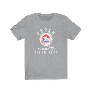 May include: A heather grey t-shirt with a white graphic design. The graphic features a red sun, a mountain range, a blue and white temple, and pink cherry blossoms. The text "JAPAN is calling AND I MUST GO" is printed below the graphic.