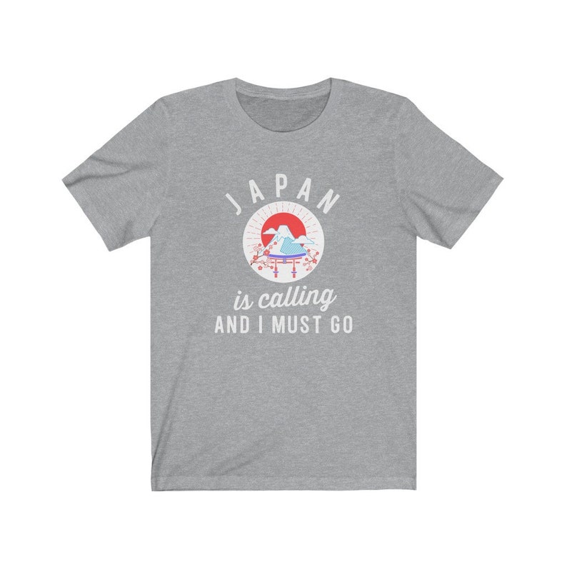 May include: A heather grey t-shirt with a white graphic design. The graphic features a red sun, a mountain range, a blue and white temple, and pink cherry blossoms. The text "JAPAN is calling AND I MUST GO" is printed below the graphic.