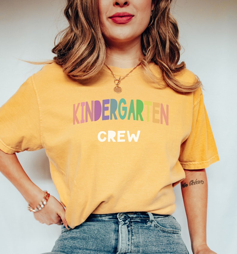 May include: A yellow t-shirt with the text "KINDERGARTEN CREW" in rainbow letters. The letters are in a rainbow gradient, with each letter a different color.