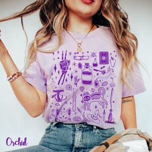 May include: Purple tie-dye t-shirt with a graphic print of various witchy symbols, including a skeleton hand, a cat, a potion bottle, a broom, and a crescent moon. The text "Orchid" is printed on the bottom left corner of the image.