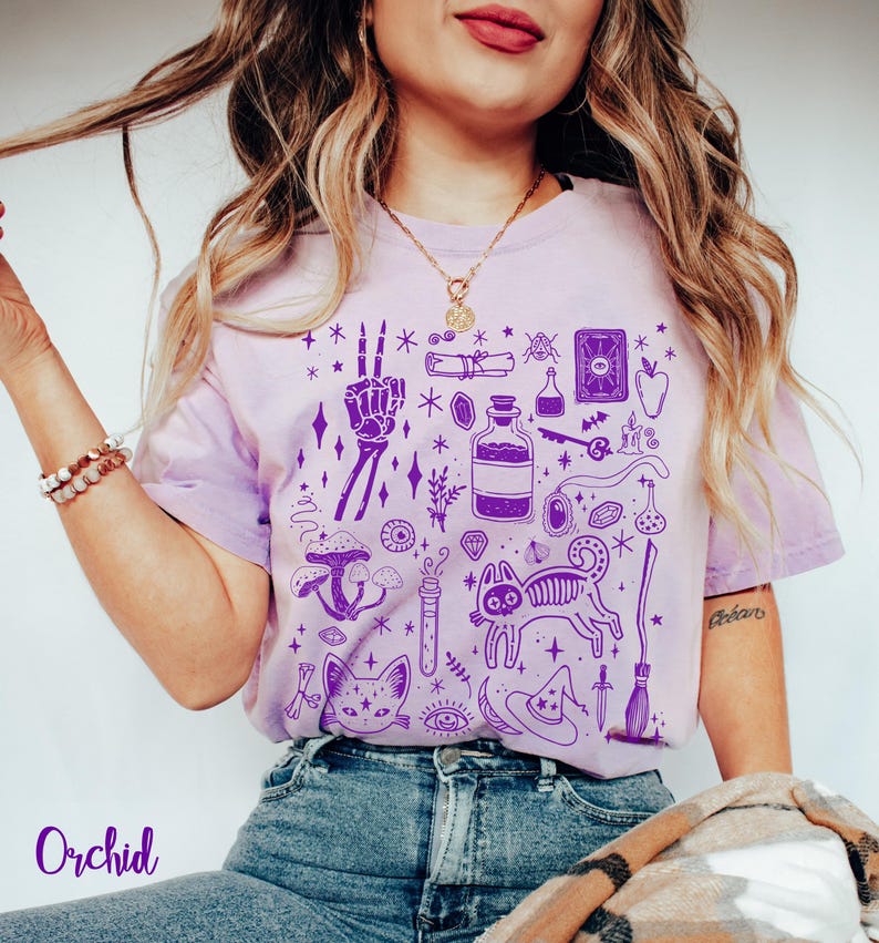 May include: Purple tie-dye t-shirt with a graphic print of various witchy symbols, including a skeleton hand, a cat, a potion bottle, a broom, and a crescent moon. The text "Orchid" is printed on the bottom left corner of the image.