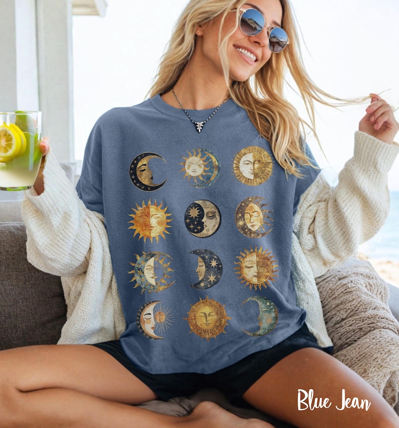 May include: A blue t-shirt with a graphic print of multiple suns and moons in gold and white. The shirt has a relaxed fit and a crew neck.