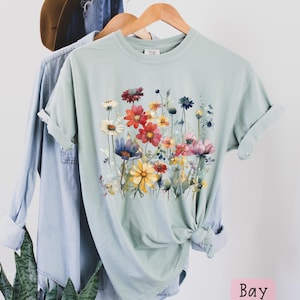 May include: A light green t-shirt with a watercolor floral print featuring a variety of colorful flowers. The flowers are arranged in a cluster and are surrounded by green leaves. The shirt has a crew neck and short sleeves.