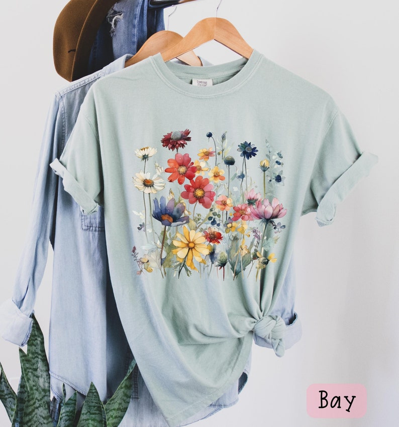 May include: A light green t-shirt with a watercolor floral print featuring a variety of colorful flowers. The flowers are arranged in a cluster and are surrounded by green leaves. The shirt has a crew neck and short sleeves.