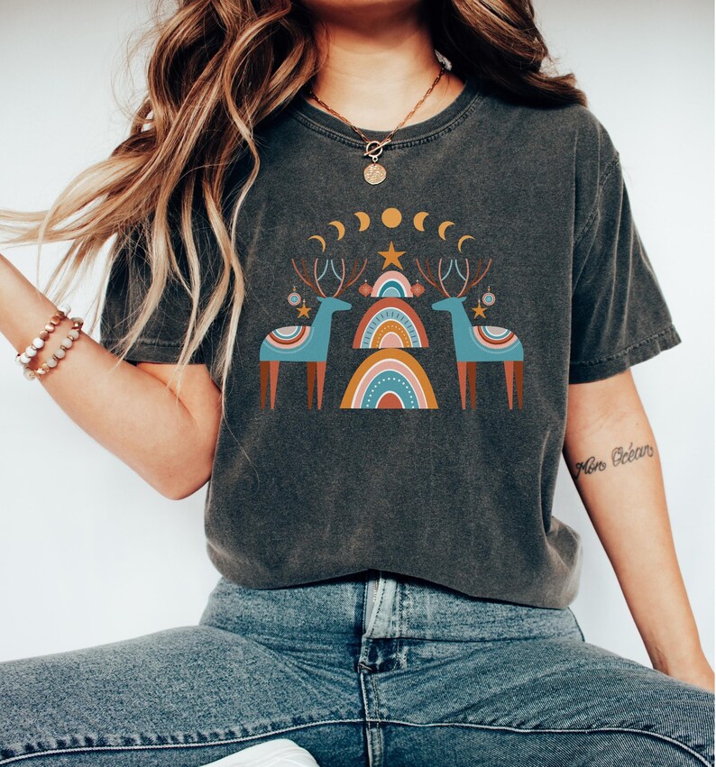 Puede incluir: Una camiseta gris oscuro con un dise&ntilde;o gr&aacute;fico que presenta dos renos estilizados de pie frente a un arco&iacute;ris. El dise&ntilde;o tambi&eacute;n incluye una estrella, lunas crecientes y peque&ntilde;os c&iacute;rculos.