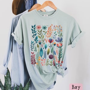 May include: A light blue t-shirt with a colorful floral print. The print features a variety of flowers, leaves, and stems in shades of pink, orange, yellow, green, blue, and purple. The shirt is folded and hanging on a wooden hanger.