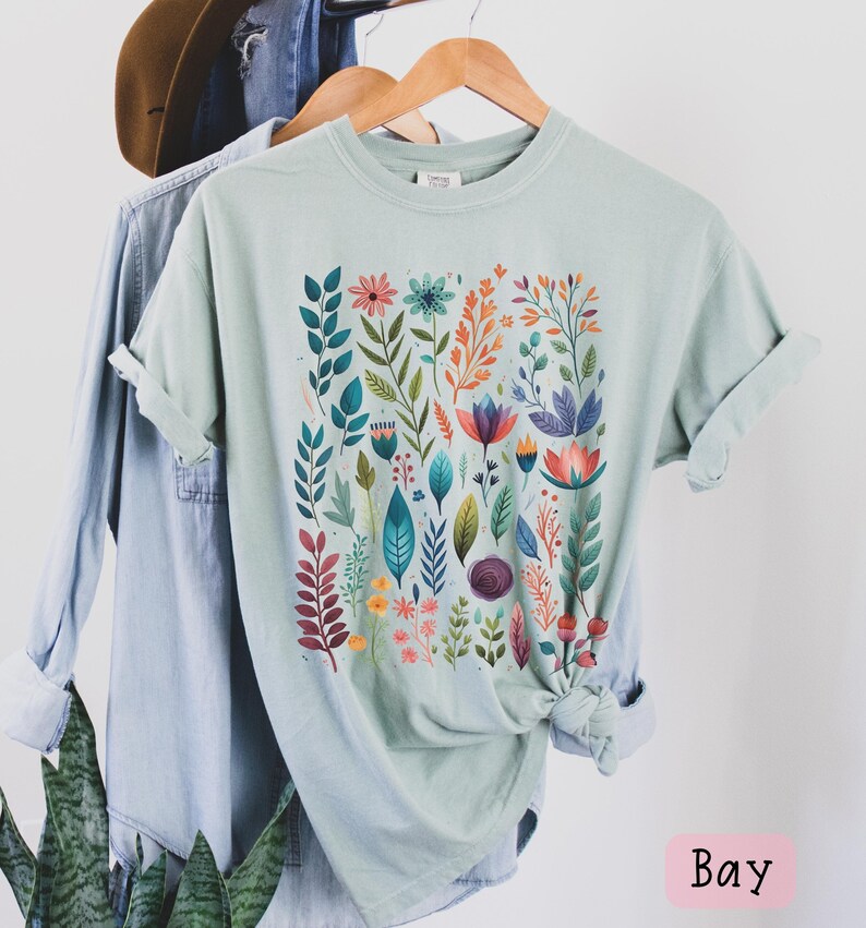 May include: A light blue t-shirt with a colorful floral print. The print features a variety of flowers, leaves, and stems in shades of pink, orange, yellow, green, blue, and purple. The shirt is folded and hanging on a wooden hanger.