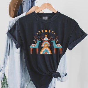 Puede incluir: Una camiseta negra con un dise&ntilde;o gr&aacute;fico de dos renos estilizados de pie frente a un arco&iacute;ris. El dise&ntilde;o tambi&eacute;n incluye una estrella, lunas y adornos.