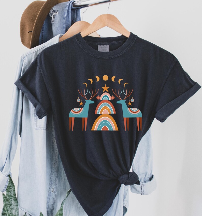 Puede incluir: Una camiseta negra con un dise&ntilde;o gr&aacute;fico de dos renos estilizados de pie frente a un arco&iacute;ris. El dise&ntilde;o tambi&eacute;n incluye una estrella, lunas y adornos.