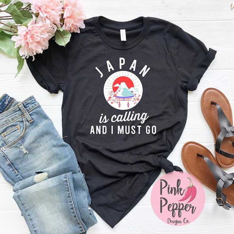 May include: Black t-shirt with a white graphic of a red sun rising over Mount Fuji with a Japanese temple and cherry blossoms. The text "JAPAN is calling AND I MUST GO" is printed below the graphic.