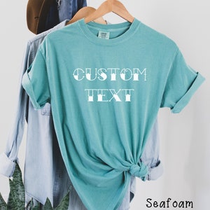 May include: A teal blue t-shirt with the text "CUSTOM TEXT" in a white, stylized font. The shirt is tied in a knot at the bottom and is hanging on a wooden hanger.
