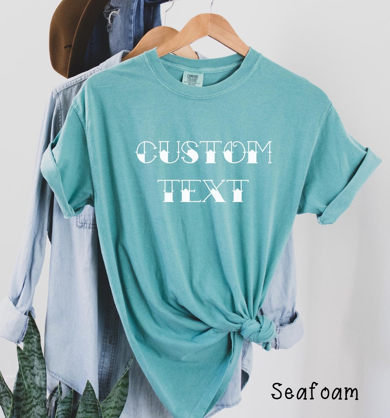 May include: A teal blue t-shirt with the text "CUSTOM TEXT" in a white, stylized font. The shirt is tied in a knot at the bottom and is hanging on a wooden hanger.