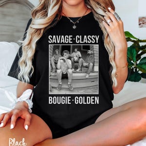Puede incluir: Camiseta negra con un gr&aacute;fico de cuatro personas sentadas en un porche. El texto "Savage. Classy. Bougie. Golden." est&aacute; impreso en la camiseta.