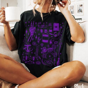 May include: Black t-shirt with a purple print of various witchy and spooky illustrations, including a cat, a skeleton hand, a potion bottle, a witch's hat, and a broom.