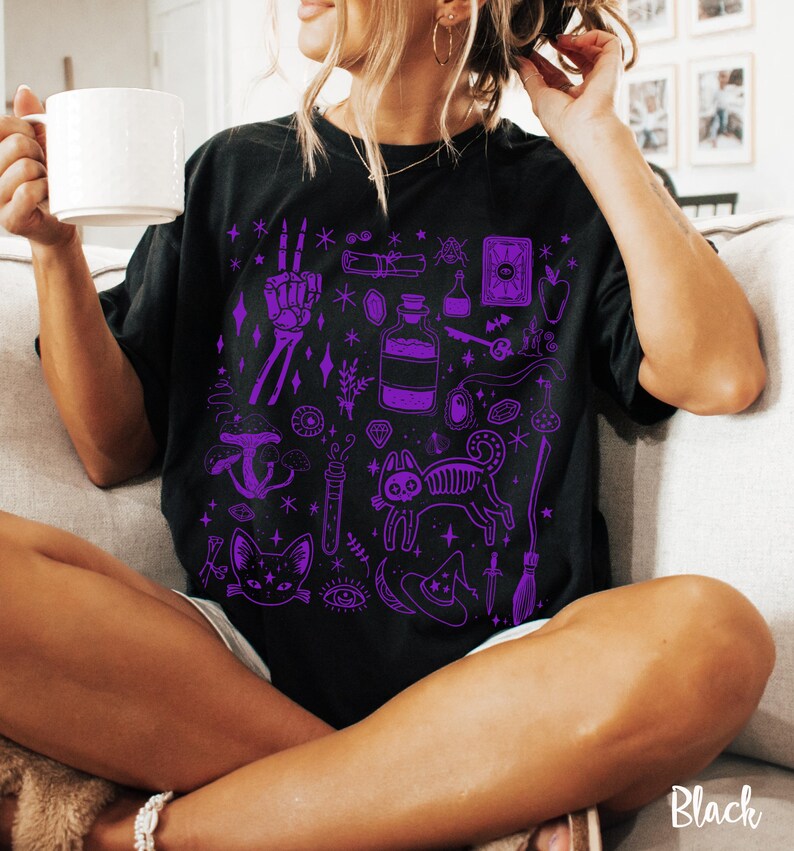 May include: Black t-shirt with a purple print of various witchy and spooky illustrations, including a cat, a skeleton hand, a potion bottle, a witch's hat, and a broom.