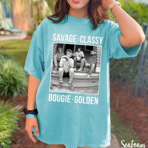 Puede incluir: Una camiseta azul turquesa con una foto en blanco y negro de cuatro personas sentadas en un porche. El texto "Savage Classy Bougie Golden" est&aacute; impreso en la camiseta.