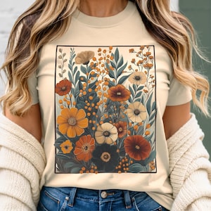 Puede incluir: Una camiseta beige con un estampado floral que presenta una variedad de flores en tonos naranja, amarillo, rojo y blanco. Las flores est&aacute;n rodeadas de hojas y tallos verdes.