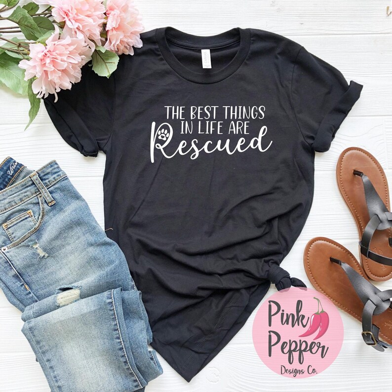 May include: Black t-shirt with white text that reads "The best things in life are Rescued" with a paw print icon. The shirt is laying on a white surface with a pair of blue jeans and brown sandals.