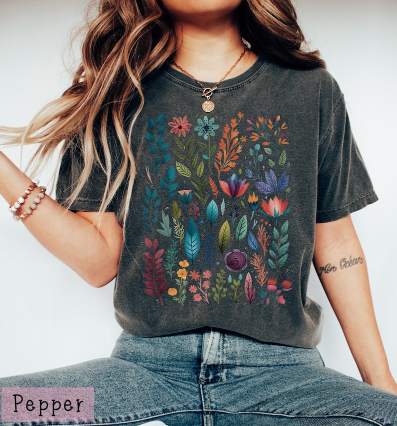 May include: A dark gray t-shirt with a colorful floral print. The print features a variety of flowers, leaves, and stems in shades of pink, blue, green, yellow, and orange.