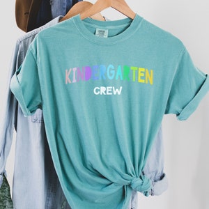 May include: A teal blue t-shirt with the words "Kindergarten Crew" in rainbow letters. The shirt is tied in a knot at the bottom.