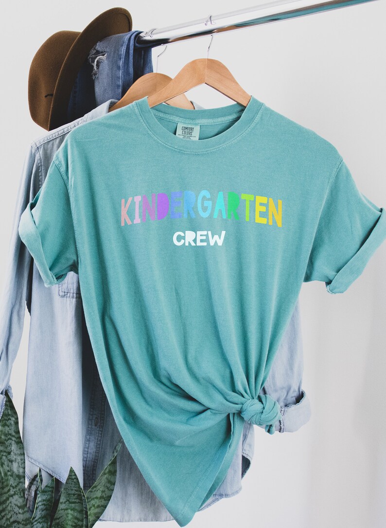May include: A teal blue t-shirt with the words "Kindergarten Crew" in rainbow letters. The shirt is tied in a knot at the bottom.