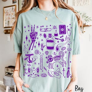 May include: A light blue t-shirt with a purple print of various witchy and mystical symbols, including a cat, a skeleton hand, a potion bottle, a crystal, a crescent moon, a star, a wand, and a tarot card. The text "Bay" is visible in the lower right corner of the image.