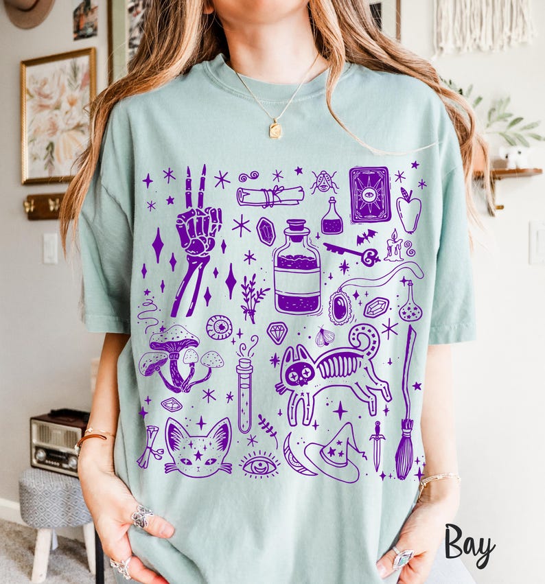 May include: A light blue t-shirt with a purple print of various witchy and mystical symbols, including a cat, a skeleton hand, a potion bottle, a crystal, a crescent moon, a star, a wand, and a tarot card. The text "Bay" is visible in the lower right corner of the image.