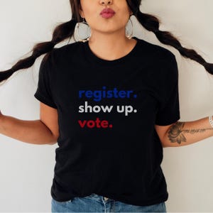 May include: A black t-shirt with the text "register. show up. vote." in blue, white, and red lettering.