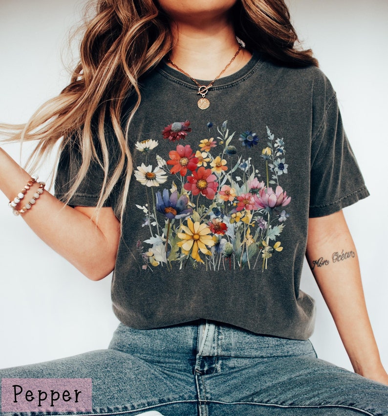 May include: A dark gray t-shirt with a watercolor floral print featuring a variety of colorful flowers. The shirt has a relaxed fit and a crew neck.
