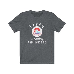 May include: A dark heather grey t-shirt with a white graphic design. The graphic features a stylized image of Mount Fuji with a red sun and cherry blossoms. The text "JAPAN is calling AND I MUST GO" is printed below the image.