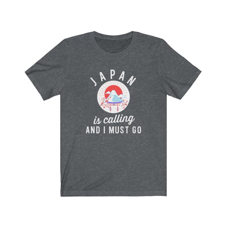 May include: A dark heather grey t-shirt with a white graphic design. The graphic features a stylized image of Mount Fuji with a red sun and cherry blossoms. The text "JAPAN is calling AND I MUST GO" is printed below the image.