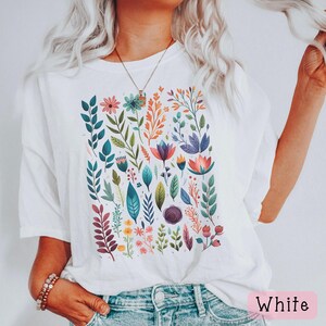 May include: White t-shirt with a colorful floral print featuring various flowers, leaves, and stems. The shirt has a relaxed fit and short sleeves.