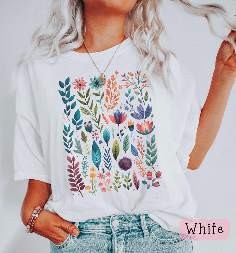May include: White t-shirt with a colorful floral print featuring various flowers, leaves, and stems. The shirt has a relaxed fit and short sleeves.