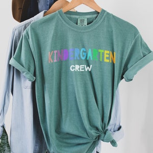 May include: A teal green t-shirt with the words "Kindergarten Crew" printed in rainbow colors. The shirt is tied in a knot and hanging on a wooden hanger.